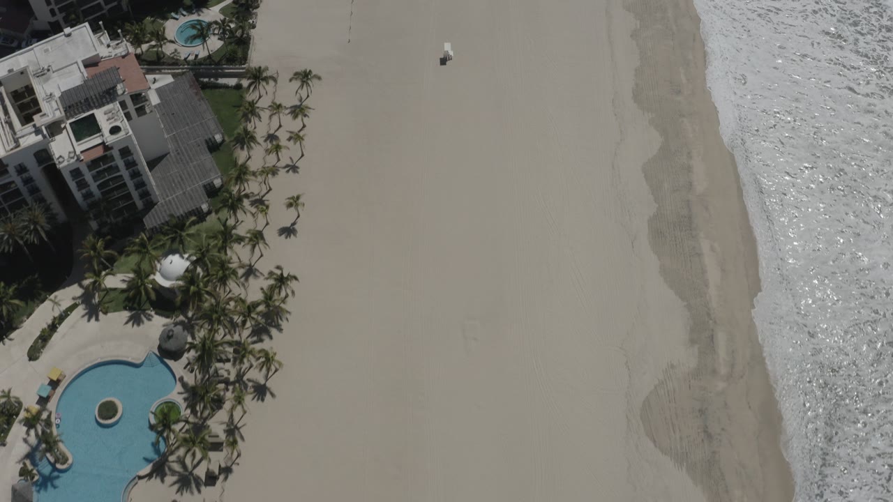 High overhead view in San Jose del Cabo showing beachfront buildings and palms at left, a blue pool below, pale sand through the center, white surf at right, and a tiny person on the beach.