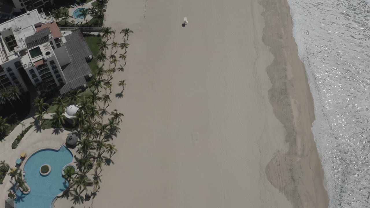 Aerial frame over San Jose del Cabo with the curving pool lower left, palms around beachfront buildings, a wide empty-looking sand field, surf foam along the right edge, and a tiny beach figure near the upper sand.