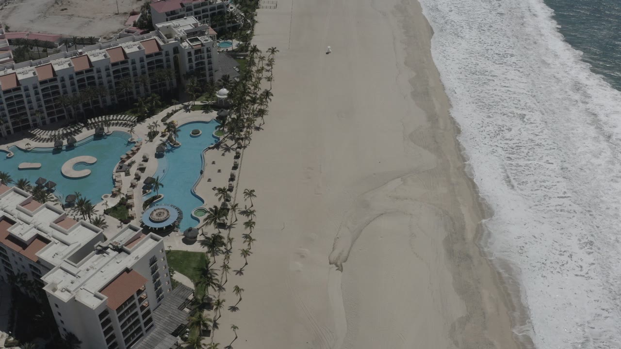 Top-down aerial view of a San Jose del Cabo resort pool complex beside a broad sandy beach and white surf, with no visible people.