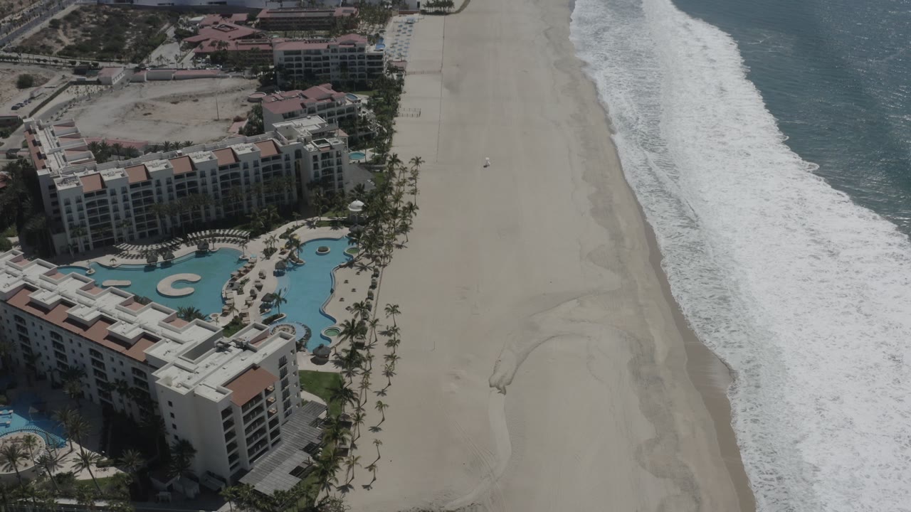 High aerial frame over San Jose del Cabo, Mexico, with the pool complex more centered at the lower edge, pale beach running upward, ocean to the right, and no visible people.