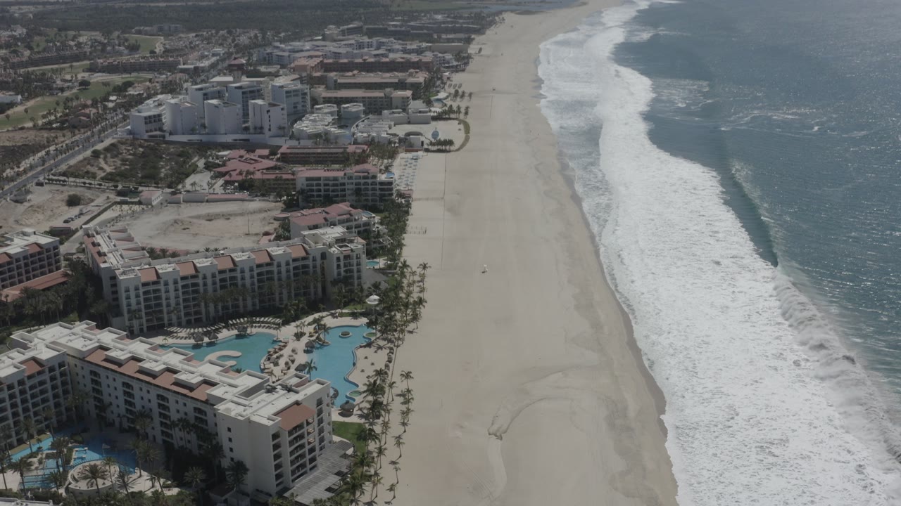 Aerial view over San Jose Del Cabo resort buildings, pools, palms, white sand beach, and blue ocean surf, with no visible people.