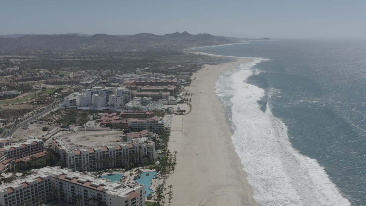 High aerial view of San Jose del Cabo resort buildings, pools, palm trees, a wide sandy beach, white surf, blue ocean, distant hills, and no visible people.