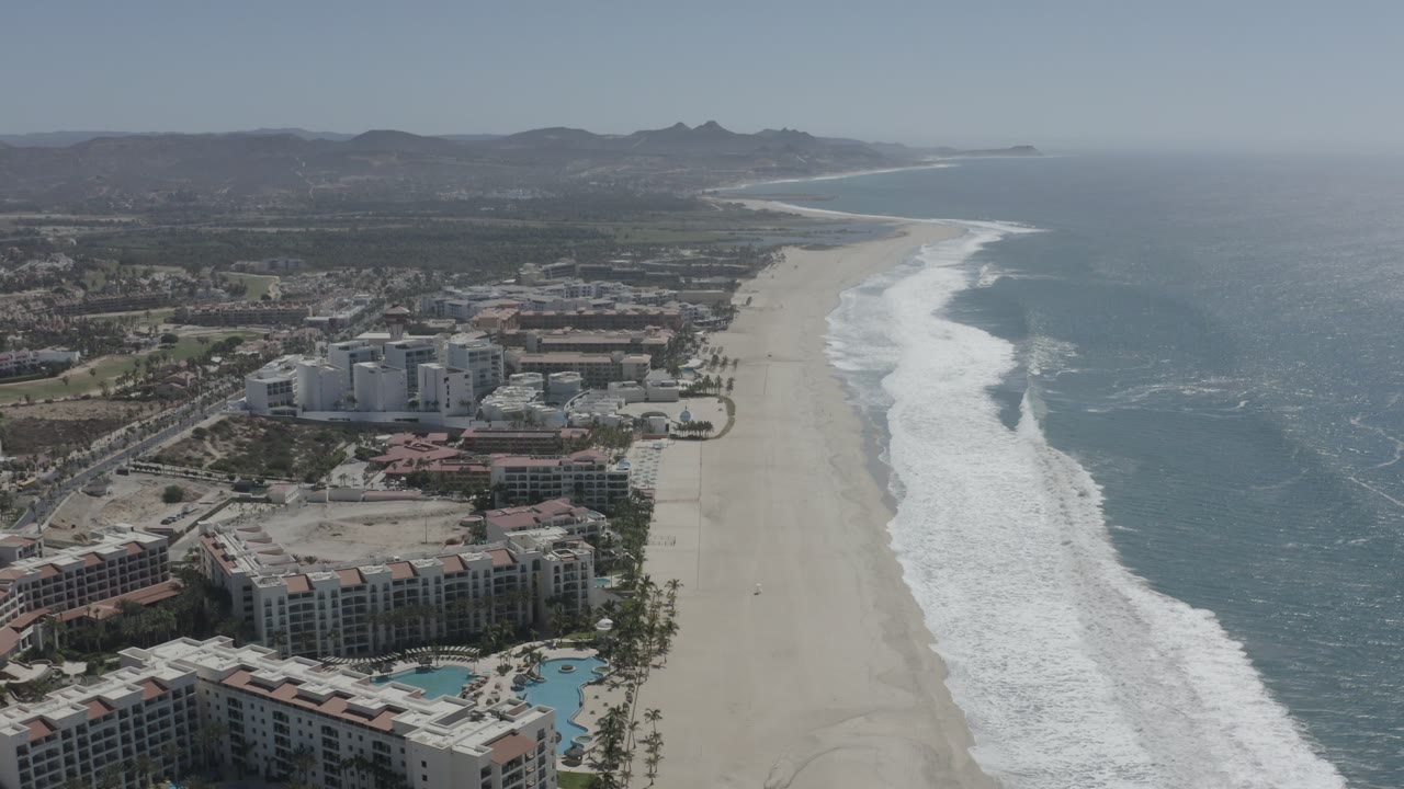 Aerial frame over San Jose del Cabo showing clustered beachfront hotels, a pale sand shoreline, foamy waves, ocean water, inland terrain, distant hills, and no visible people.