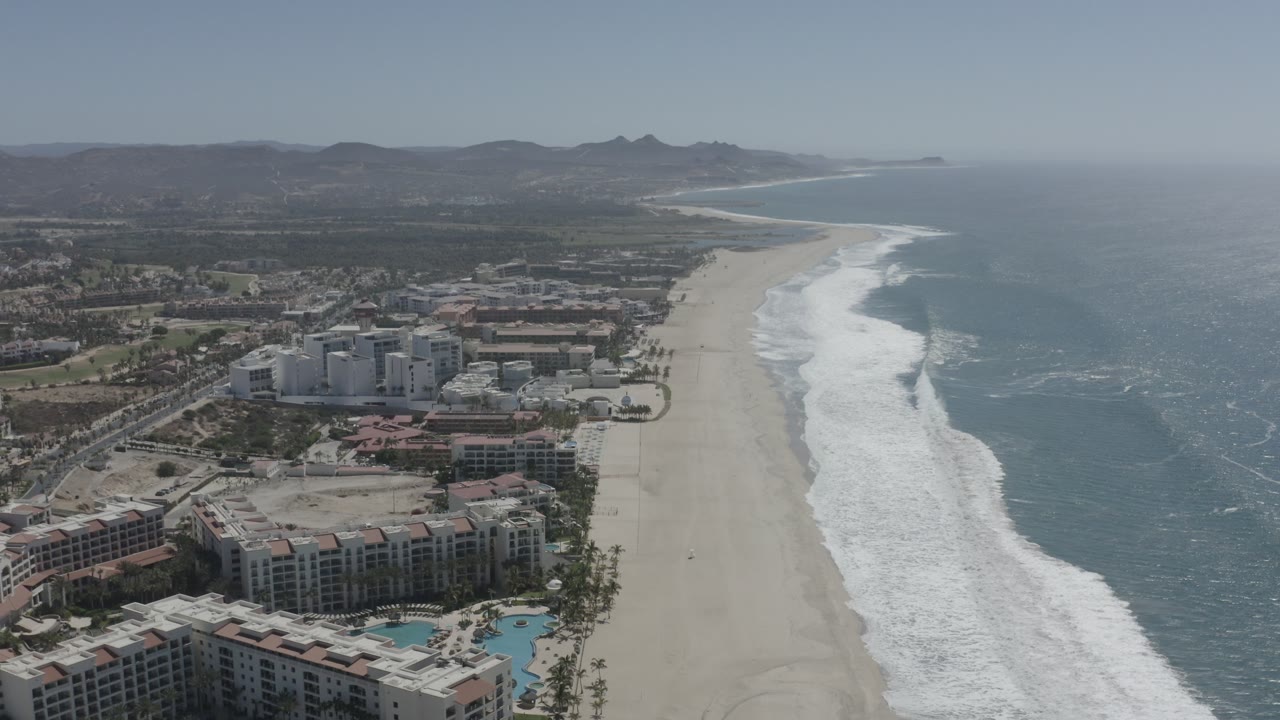 Wide beach, ocean surf, resort towers, and hills from a drone view.