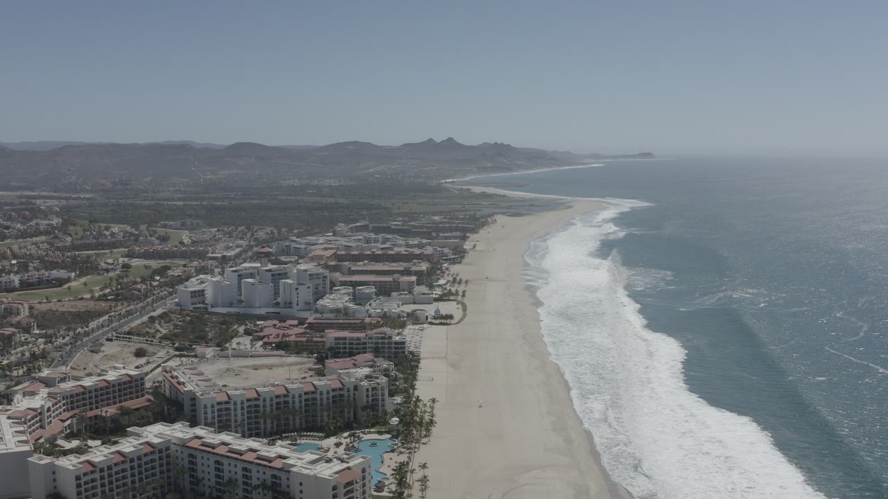 Aerial coastline with beachfront buildings, sand beach, surf, ocean, and distant hills.