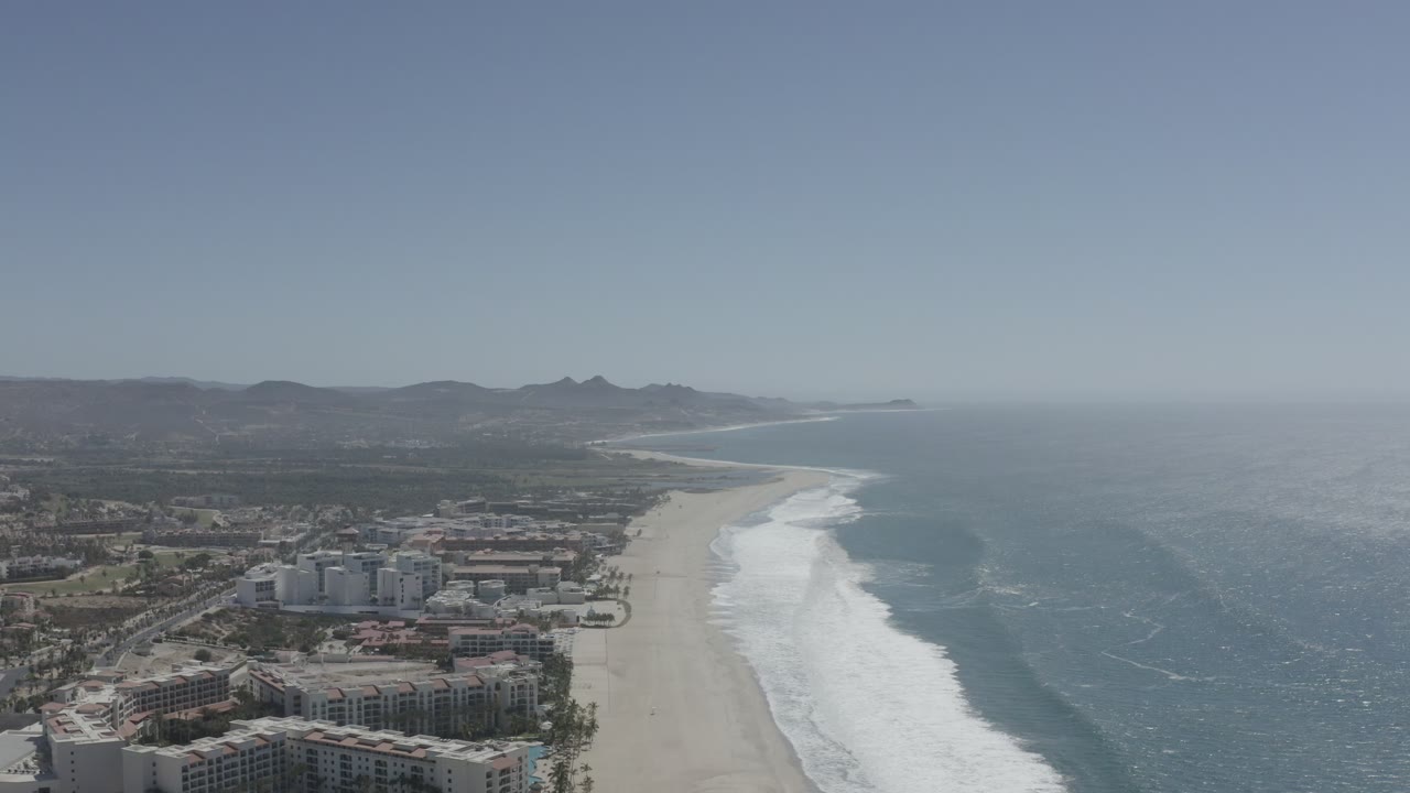 Aerial coastline with beachfront buildings, sandy beach, surf, ocean, distant hills, and blue sky.