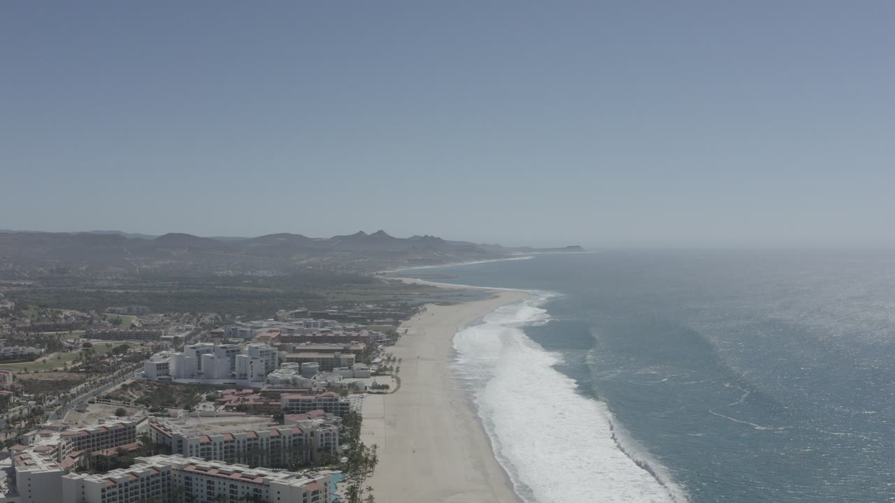 Aerial view of beachfront buildings, sand beach, surf, ocean, and distant mountains.