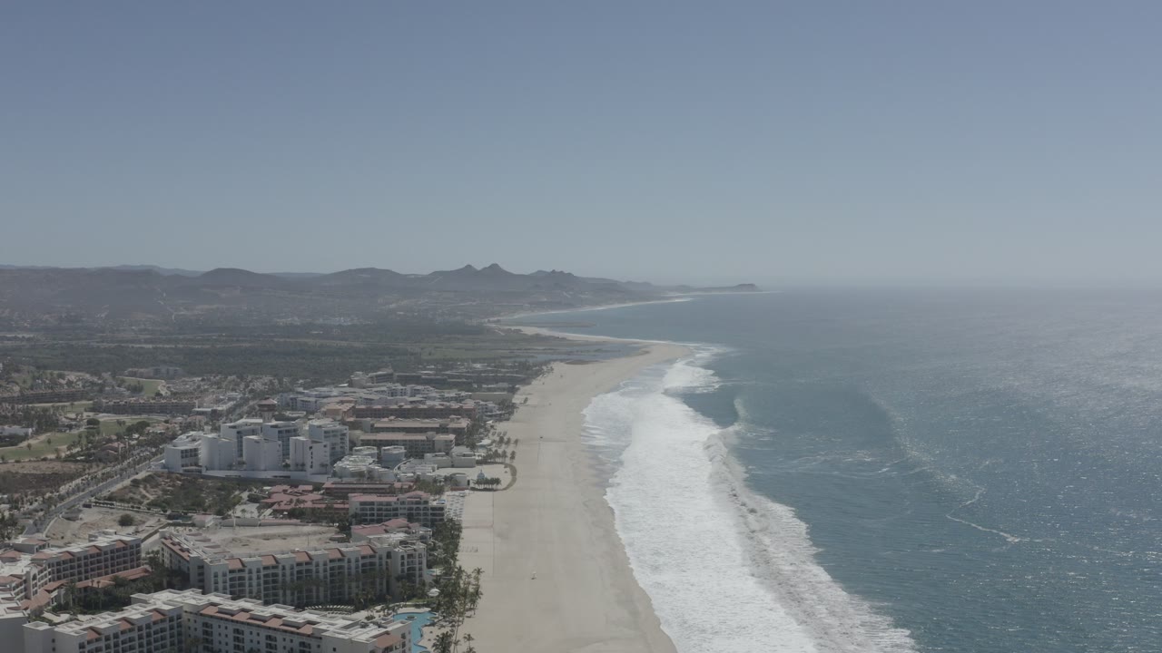 High aerial view over San Jose del Cabo with beachfront buildings at lower left, a long sandy beach, white surf, blue ocean, hazy hills, and no visible people.