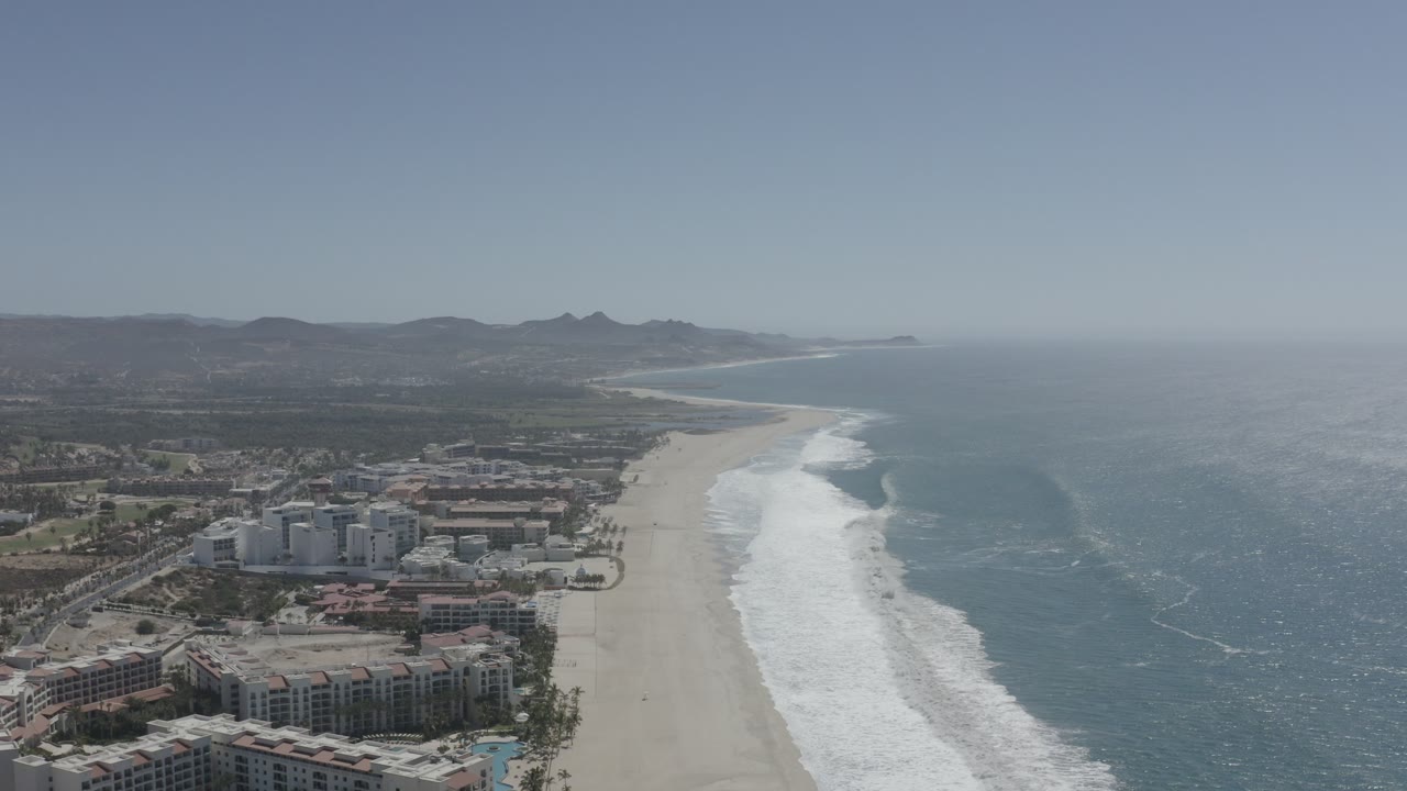 Drone view of beachfront buildings, wide sand, white surf, ocean, and hills.