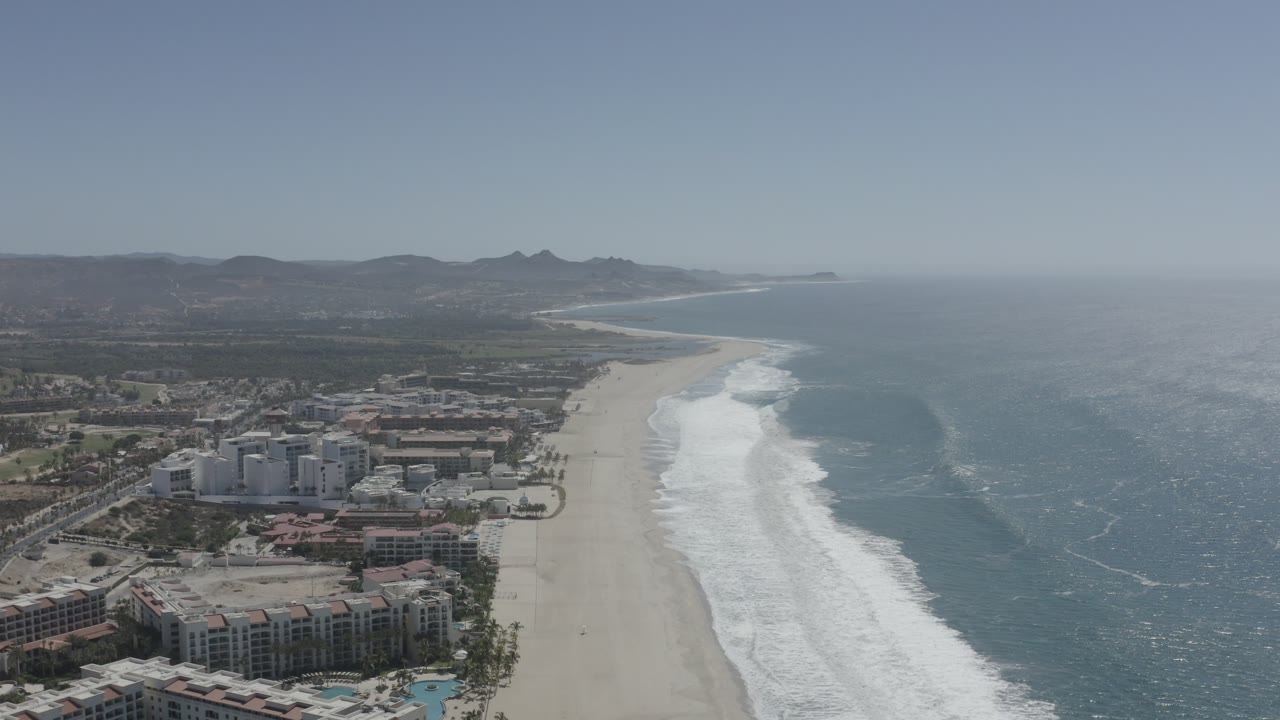 Wide aerial frame in San Jose del Cabo with resort-style buildings at the bottom edge, empty sand, long bands of breaking waves, blue water, hazy sky, and no visible people.