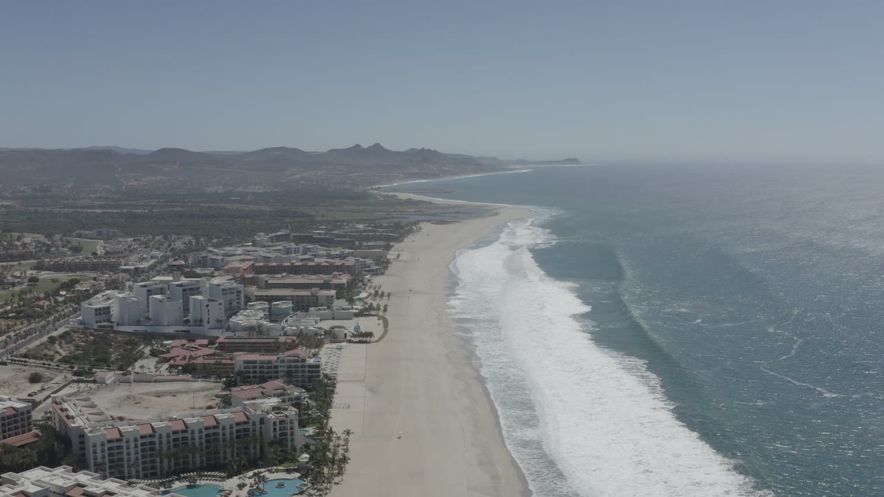 Aerial frame in San Jose del Cabo showing beachfront blocks in the foreground, the pale sand running up the coast, breaking white waves, open water, distant hills, and no visible people.