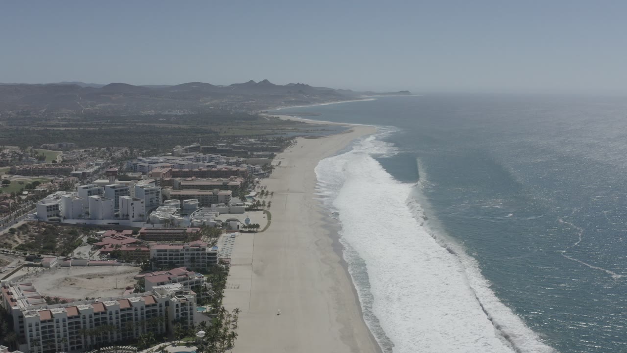 Aerial view over San Jose del Cabo with resort buildings at lower left, a broad sandy beach through the center, teal ocean to the right, and no visible people at this scale.