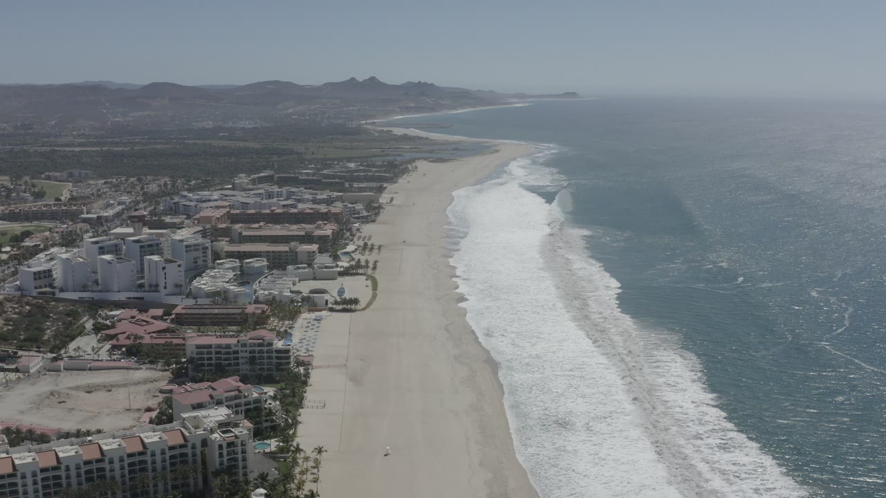 A high aerial frame over San Jose del Cabo shows resort buildings at left, a wide sandy beach through the center, blue-green ocean at right, distant hills, hazy daylight, and no visible people.