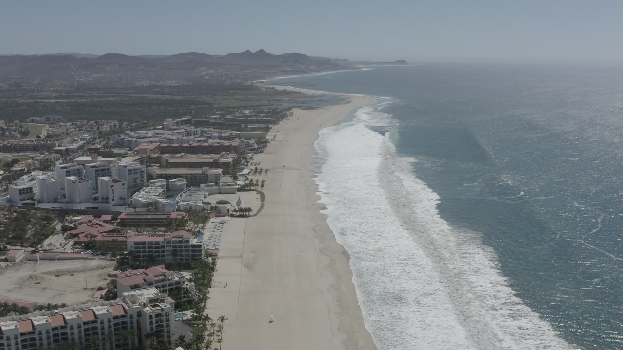 Aerial coastline view with ocean surf, beach, resort area, and mountains beyond.