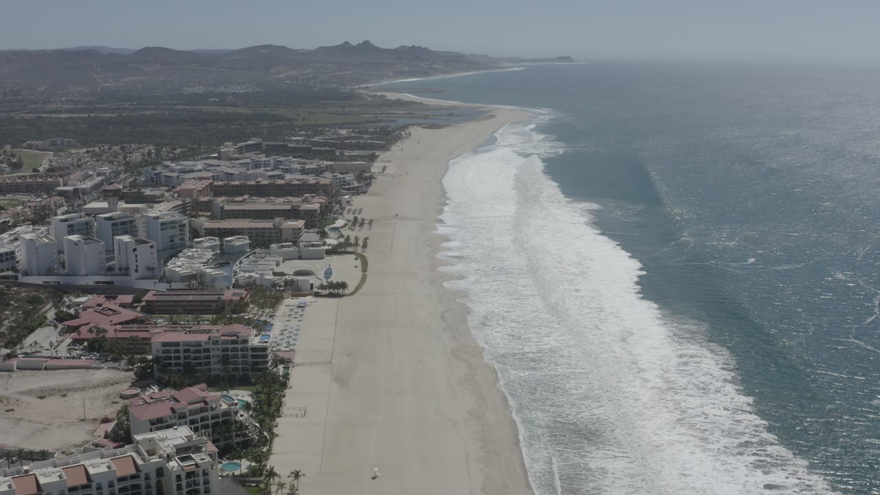 Aerial frame over San Jose del Cabo showing beachfront buildings at lower left, a wide pale beach, white surf bands, blue-green ocean, distant hills, and no visible people.