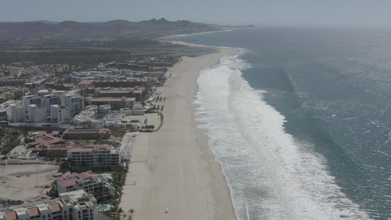 Aerial view in San Jose del Cabo with resort-scale buildings nearer the lower left edge, long sand beach through the middle, bright surf to the right, hazy coastline, and no visible people.