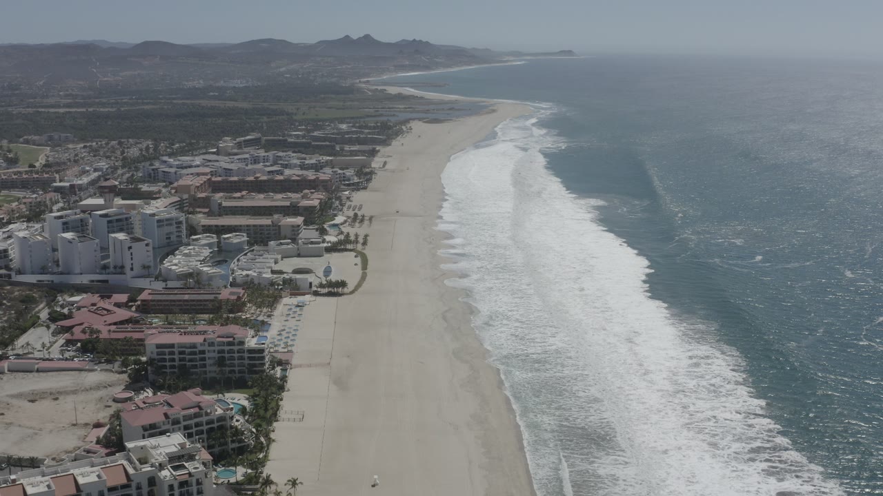 Aerial shoreline with white surf, open ocean, beach buildings, and mountain backdrop.