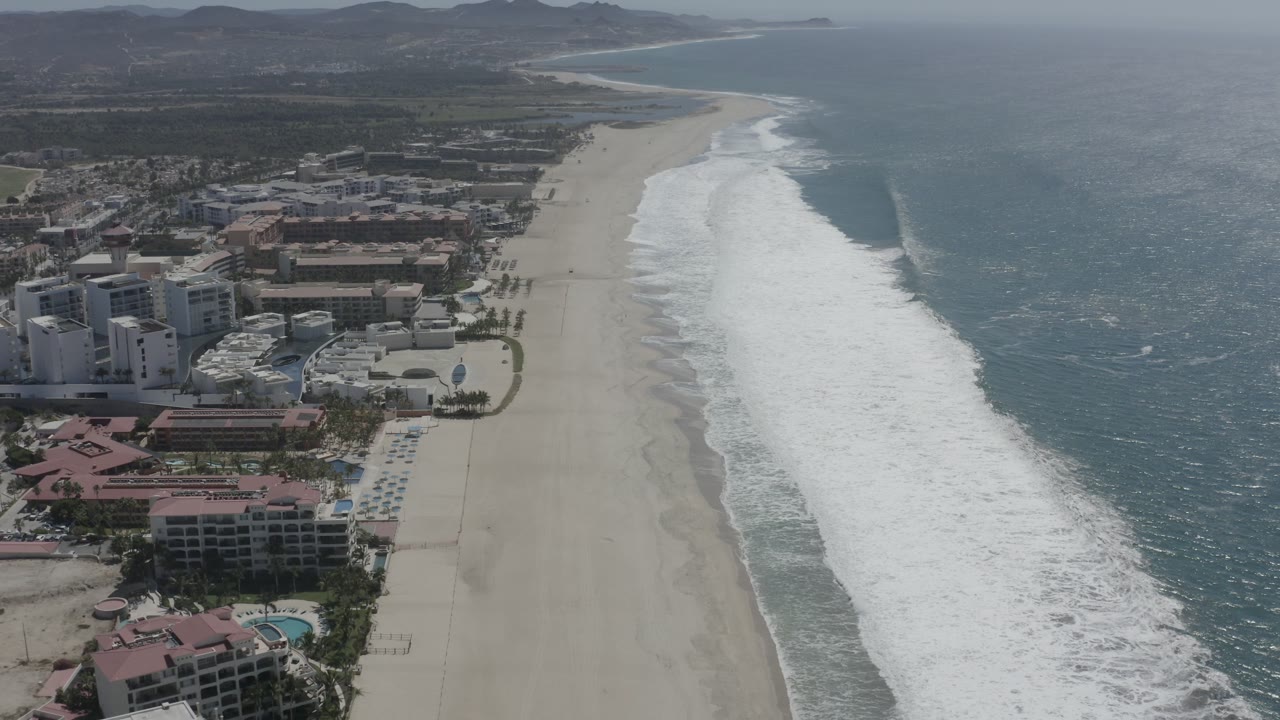 Mid-clip aerial view in San Jose Del Cabo, Mexico, keeping the resort strip on the left, the beach as a vertical band, and darker blue ocean water filling the right side.