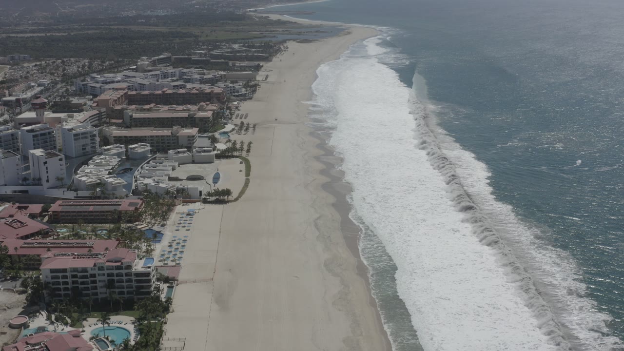 Opening aerial frame over San Jose del Cabo, Mexico, with beachfront buildings and pools at left, a wide pale beach in the center, blue ocean at right, and no visible people.