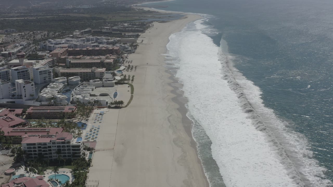 Aerial frame over San Jose del Cabo showing the shoreline slightly farther along, with red-roofed beachfront buildings, open sand, foamy surf, blue water, and no visible people.
