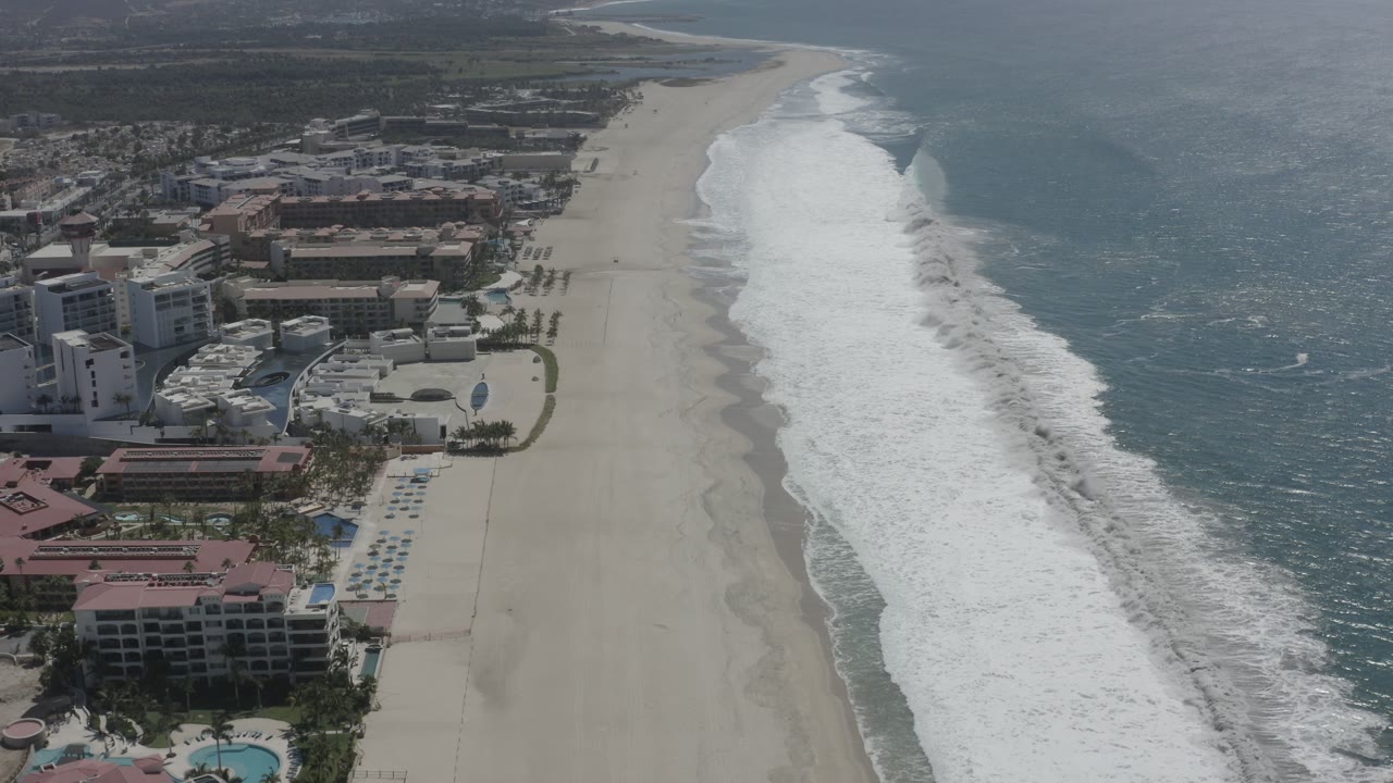 Aerial shoreline view with hotels, beach, foam bands, and ocean water.