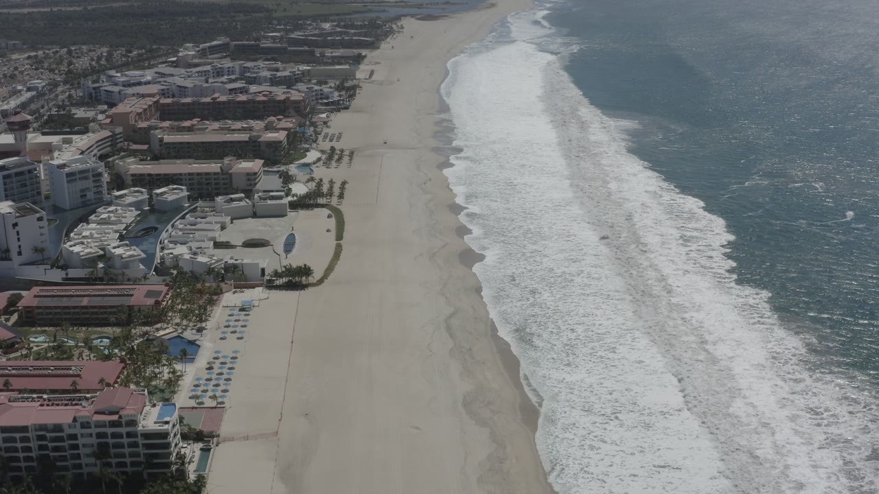 High aerial frame in San Jose del Cabo, Mexico, showing white wave foam moving diagonally along the beach, with resort roofs, pools, palms, and small beach figures near the left edge.