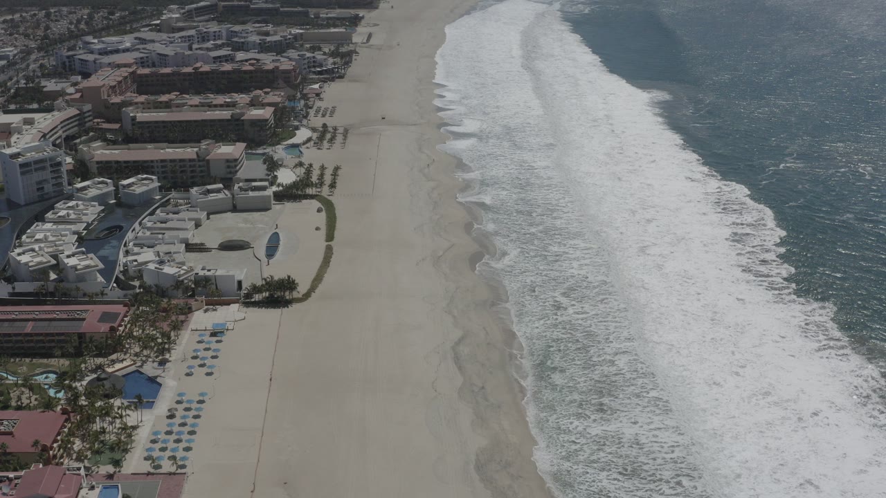 High aerial view over San Jose Del Cabo, Mexico, with resort buildings at left, pale sand through the center, blue ocean at right, and no visible people.