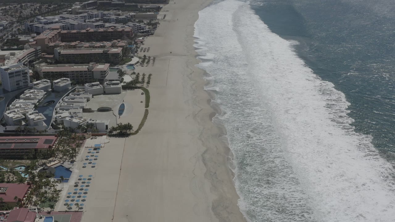 Aerial frame in San Jose Del Cabo showing a diagonal strip of foamy surf beside a wide beach, with resort structures and palm landscaping on the left and no visible people.