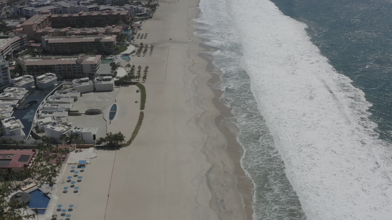 Aerial view in San Jose Del Cabo showing the surf band farther along the right edge, open sand in the center, resort-style pools below, and no visible people.