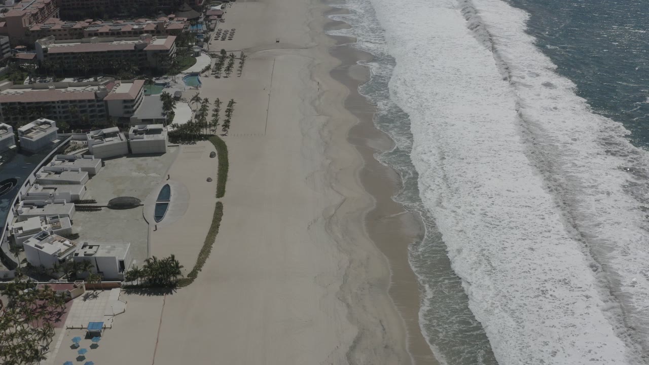 Aerial view of resort buildings beside a sandy beach and foaming surf.