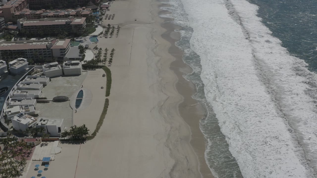Aerial frame over San Jose del Cabo, Mexico, with the surf shifted along the right side, resort pools and white buildings on the left, and no visible people on the sand.