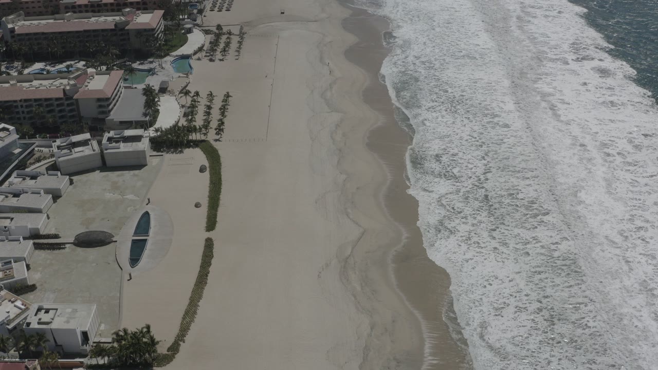 Aerial view of beach surf beside beachfront buildings and pools.