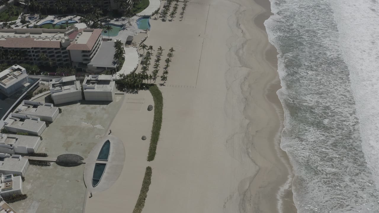 High aerial view in San Jose Del Cabo with resort-like buildings on the left, open sand in the middle, and breaking surf along the right, with no visible people.