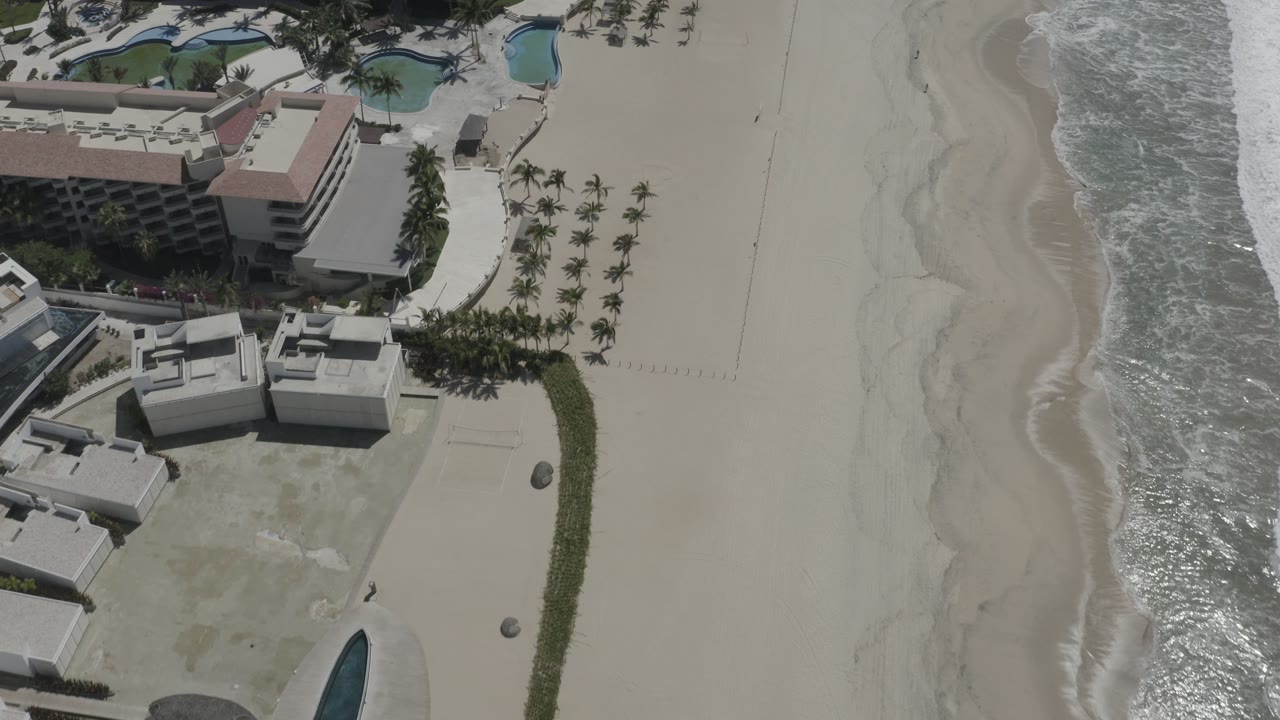 Aerial frame in San Jose del Cabo with a resort-style building, blue pools, palms, empty sand, and bright reflected surf, with no visible people.