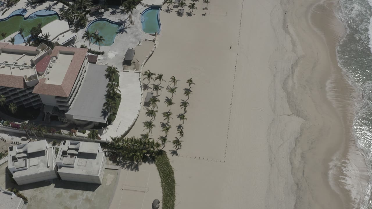 Overhead view in San Jose Del Cabo, Mexico showing resort roofs, palm-lined grounds, curved pools, wide sand beach, and surf at the right edge, with no visible people.