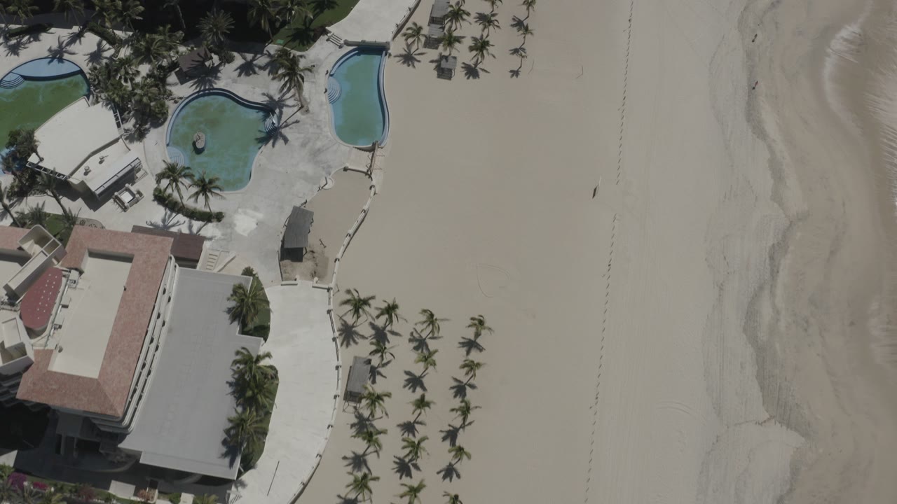 Overhead view in San Jose Del Cabo showing resort buildings, two blue pools, rows of palms, broad pale sand, shoreline texture, and no visible people.