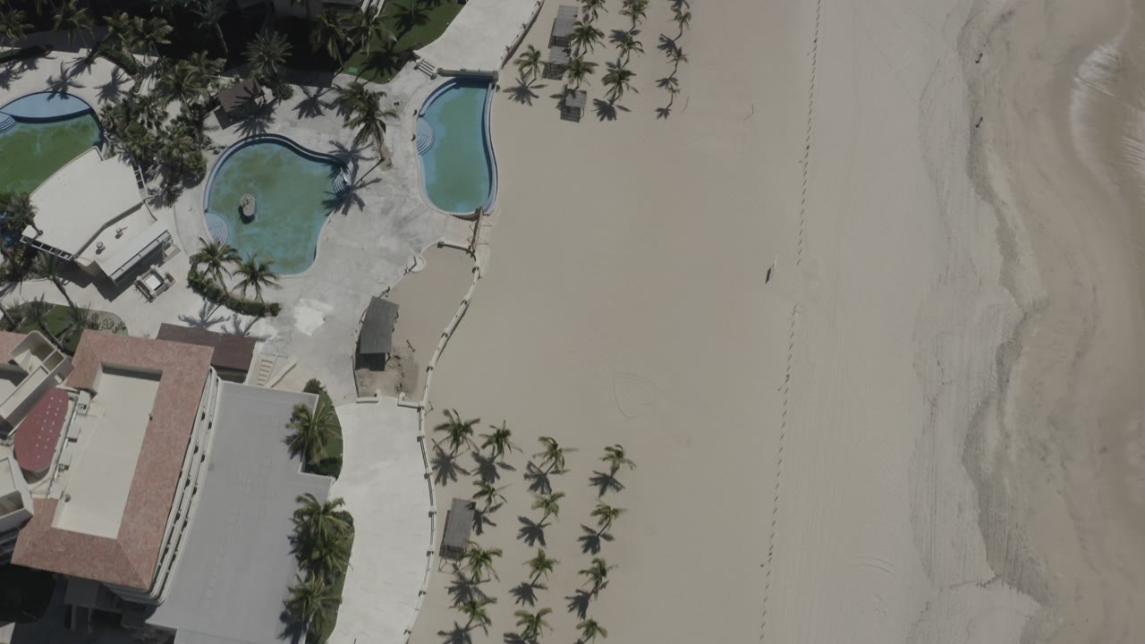 Drone view of resort grounds, palms, pools, and empty beach.