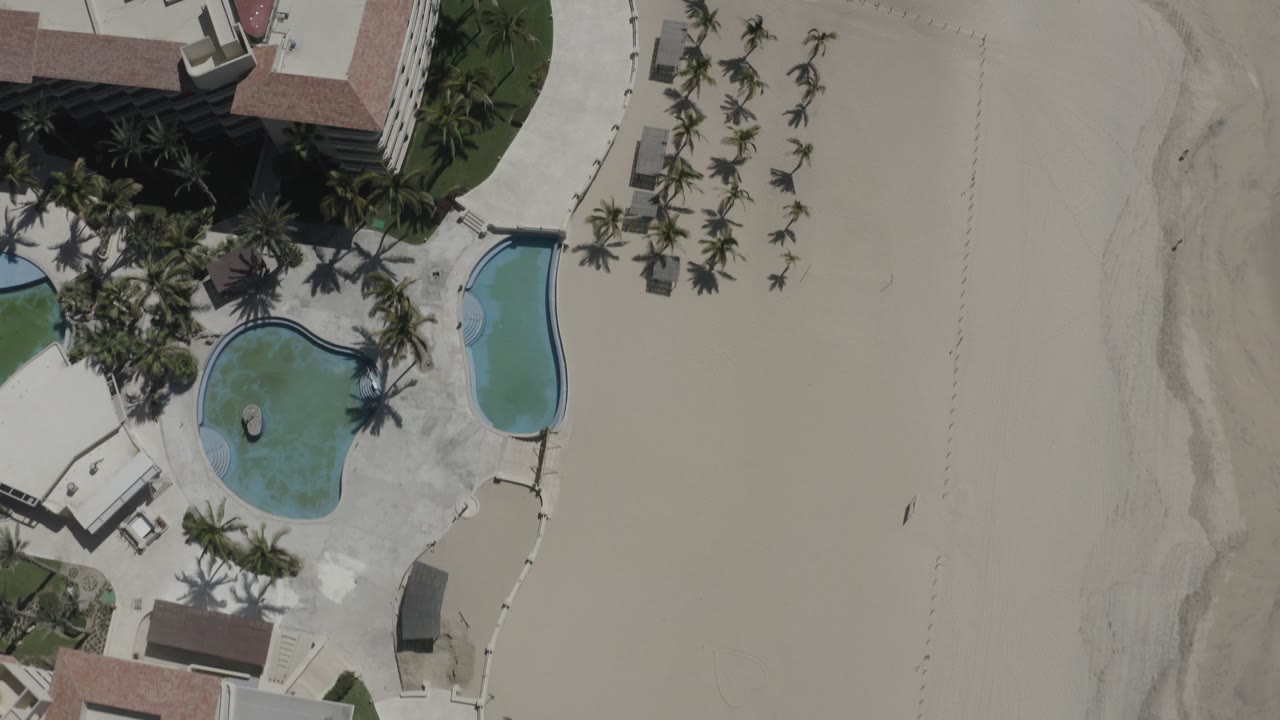 Overhead frame of a San Jose del Cabo resort with curved blue pools, palm clusters, hotel roof edges, pale sand, and tiny distant figures on the beach.