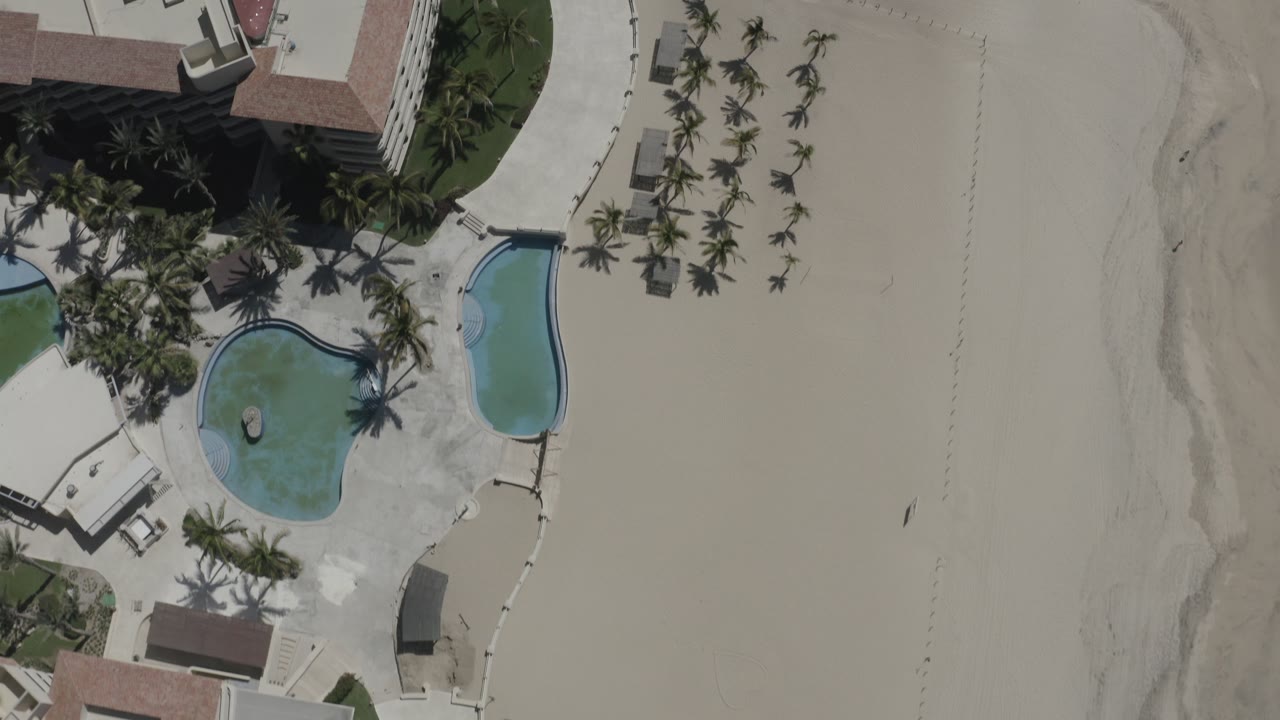 A second overhead view in San Jose del Cabo shows the hotel corner, two resort pools, palm shadows, a wide sandy beach, and small distant beach figures.