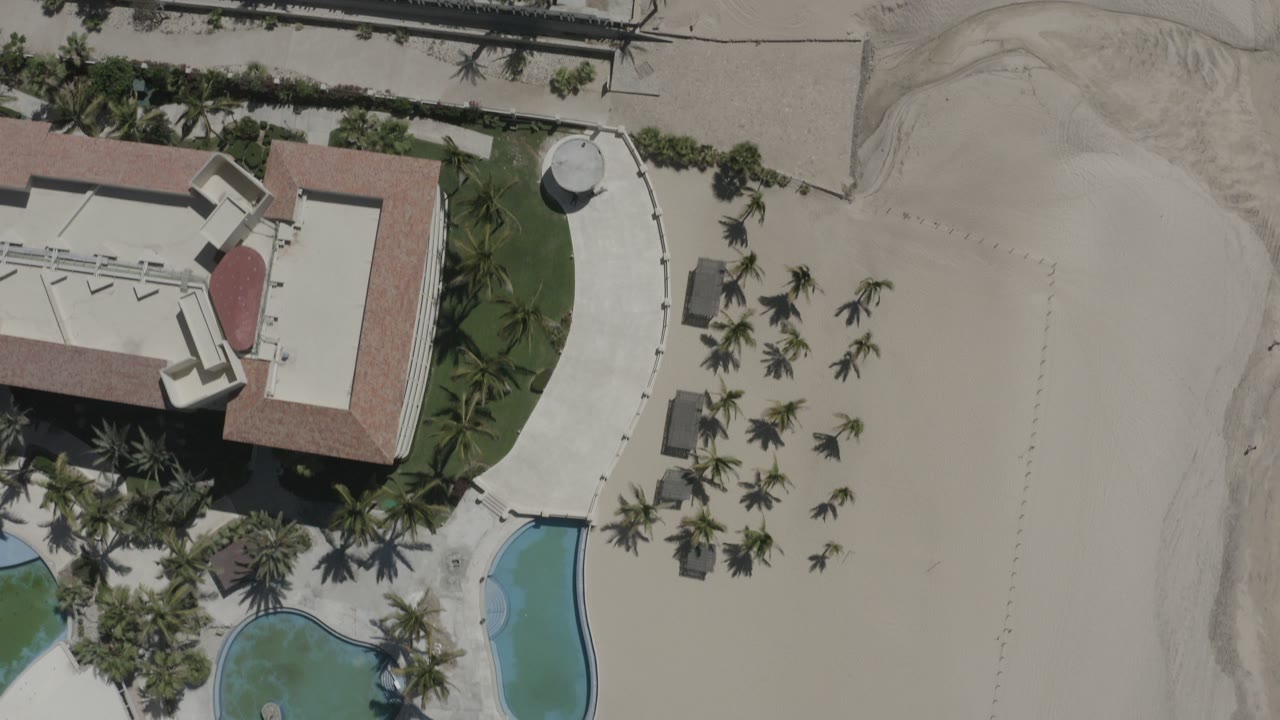 Top-down aerial frame in San Jose del Cabo, Mexico, showing resort buildings at left, curved pool water, palm landscaping, and pale beach sand with no visible people.