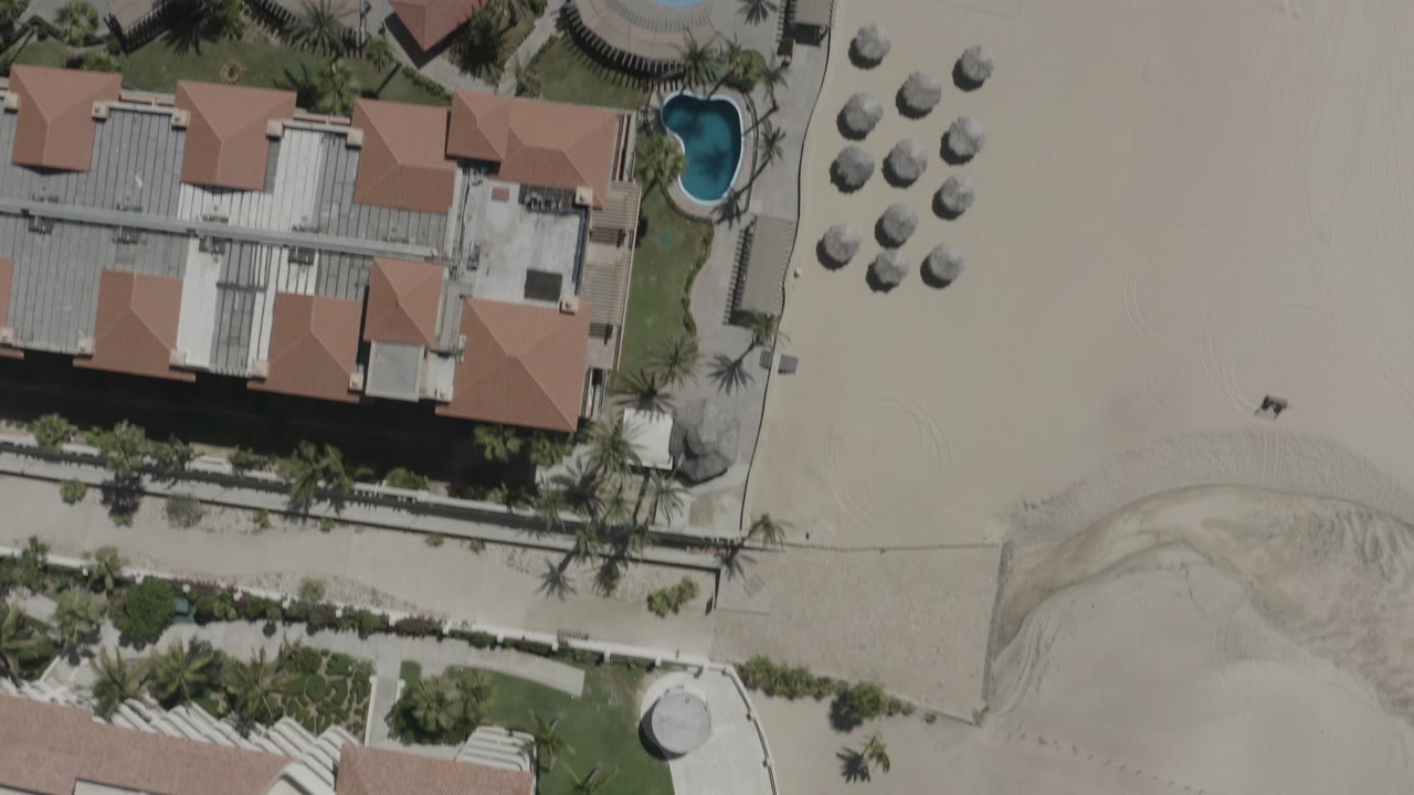 Overhead view in San Jose del Cabo, Mexico, showing terracotta-roofed resort buildings, a small blue pool, beach umbrellas, pale sand, and tiny beachgoers visible.