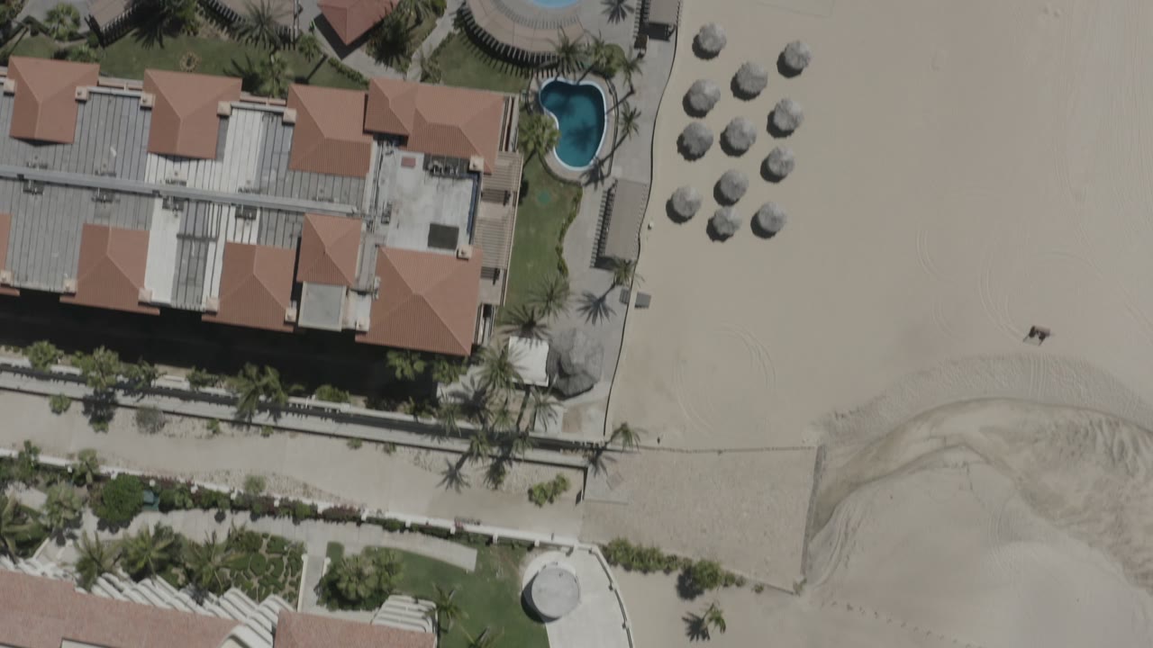 High overhead frame in San Jose del Cabo, Mexico, with hotel roofs centered, a small pool near palm landscaping, grouped umbrellas on the sand, and tiny people visible on the beach.