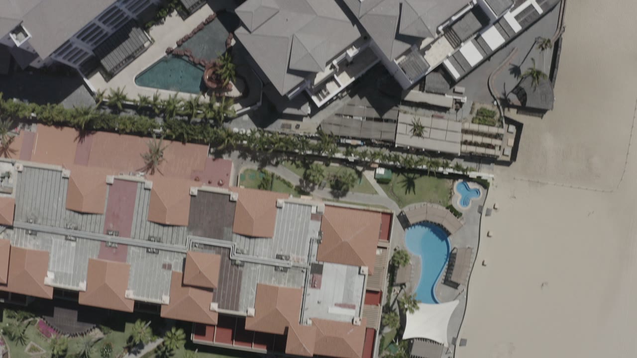 Overhead view in San Jose Del Cabo, Mexico, showing condominium roofs, two blue pools, palm landscaping, and pale beach sand with no visible people.
