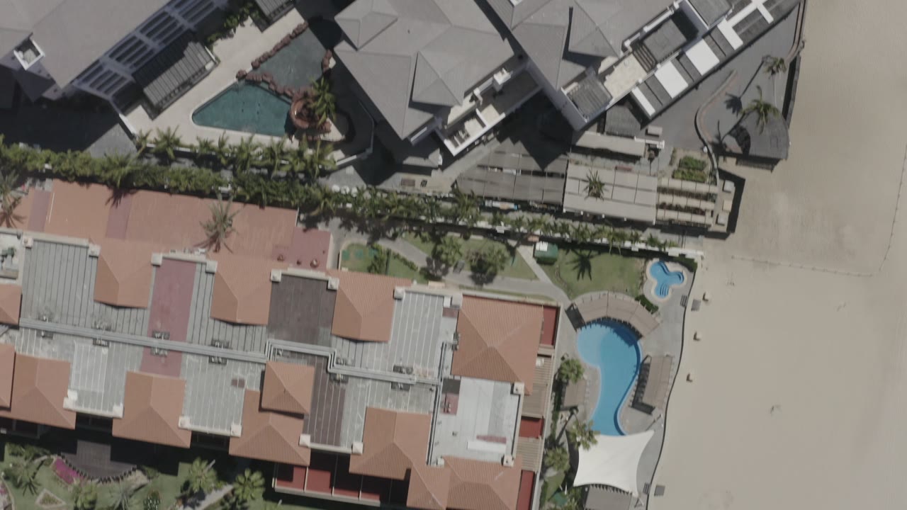 Aerial overhead frame in San Jose Del Cabo, Mexico, with light rooftops, curved pools, a palm-lined boundary, and open sand with no visible people.