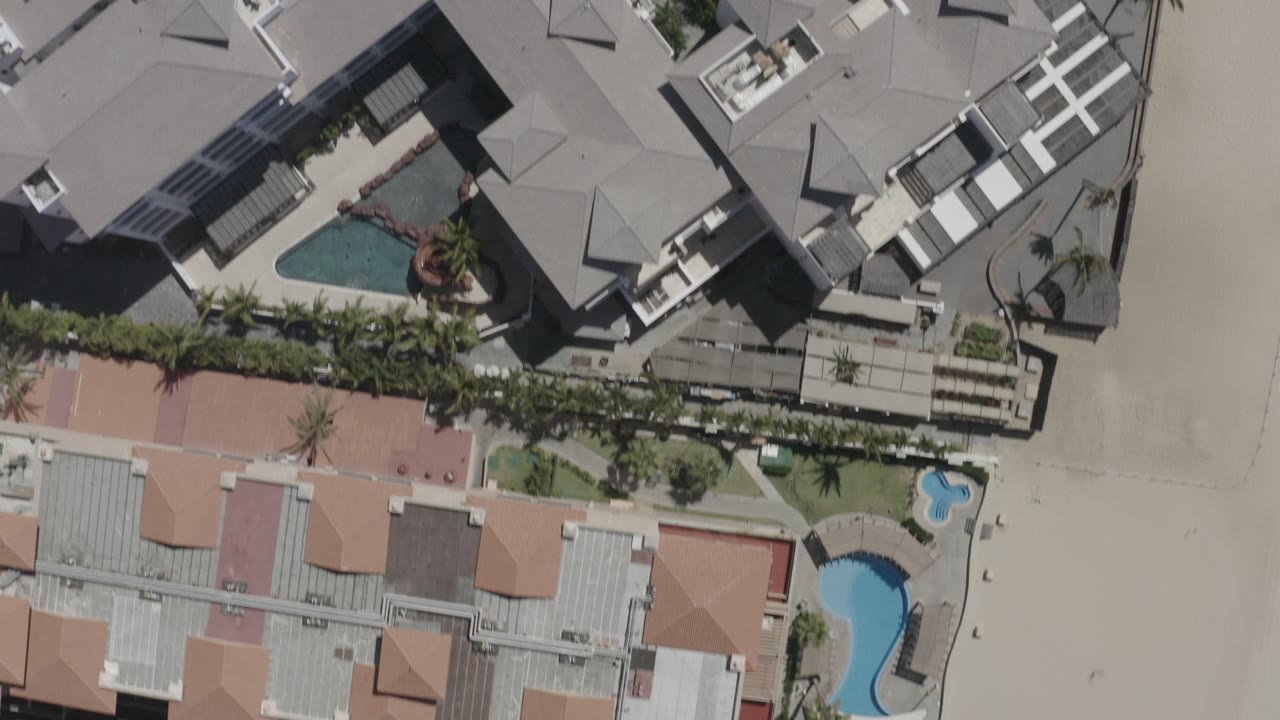 Top-down San Jose Del Cabo view showing condominium buildings, a larger blue pool, a small curved pool, palm landscaping, and beach sand with no visible people.