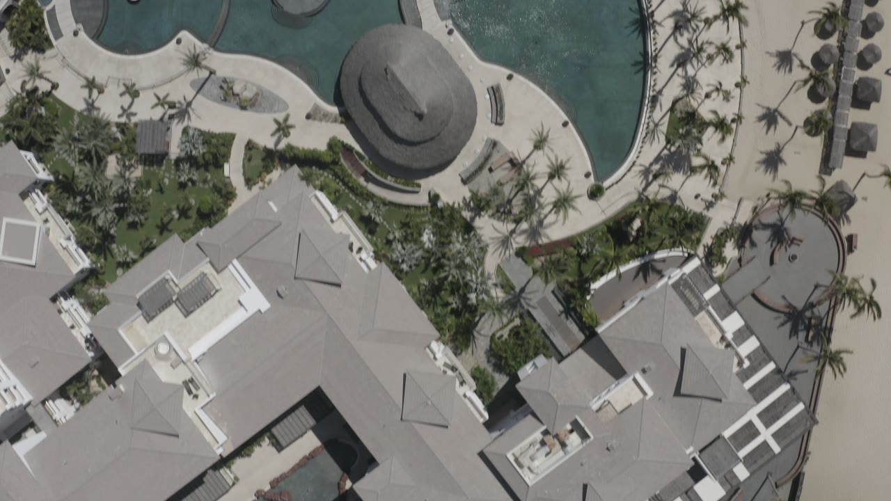 Overhead frame in San Jose del Cabo, Mexico showing curved swimming pools, gray rooftops, palm landscaping, pale concrete walkways, and no visible people.