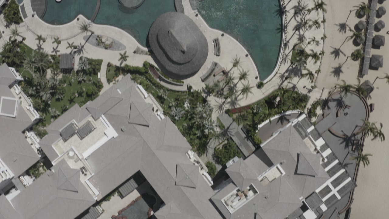 Aerial frame in San Jose del Cabo, Mexico with a large gray roof in the lower half, turquoise pools above it, palm shadows, concrete paths, and no visible people.