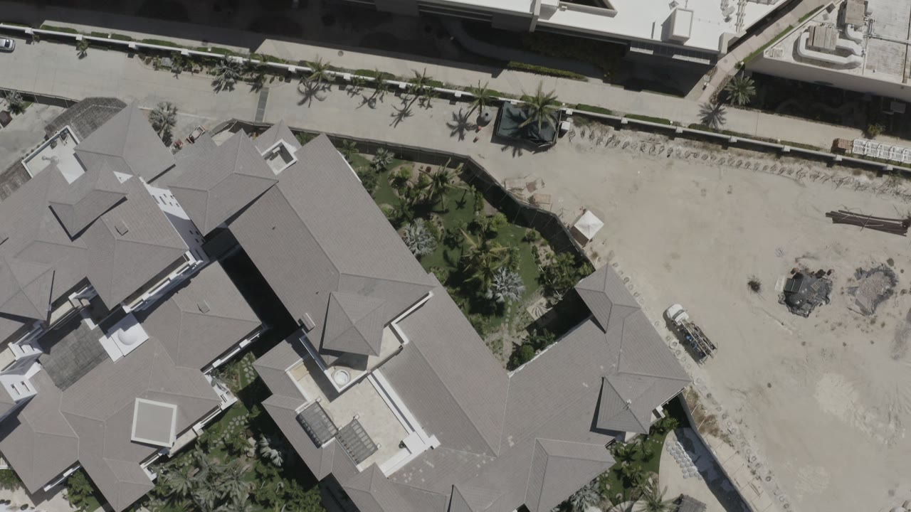 Overhead drone frame in San Jose del Cabo showing gray-roofed private homes, sandy open ground, a road edge, palms, and no visible people.