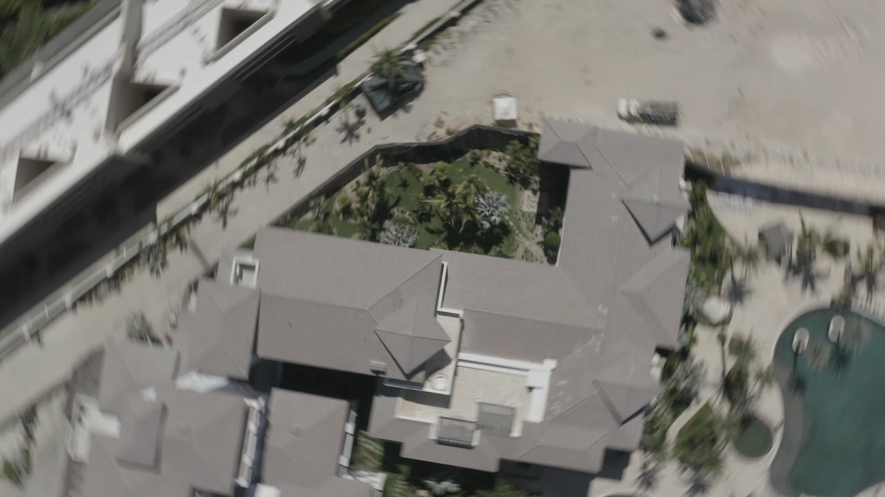 Aerial view over San Jose Del Cabo, Mexico with the mansion roof filling the lower frame, landscaping at center, a pool at right, and no visible people.