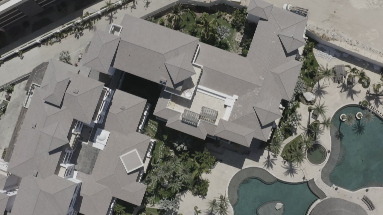 Overhead San Jose del Cabo resort frame with gray roof planes at the edges, palms around the courtyard, and connected green-blue pool shapes with no visible people.